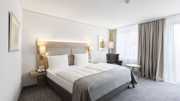Una habitación de hotel con una cama grande, mesitas de noche, una silla, una pequeña mesa redonda y un ventanal con cortinas.