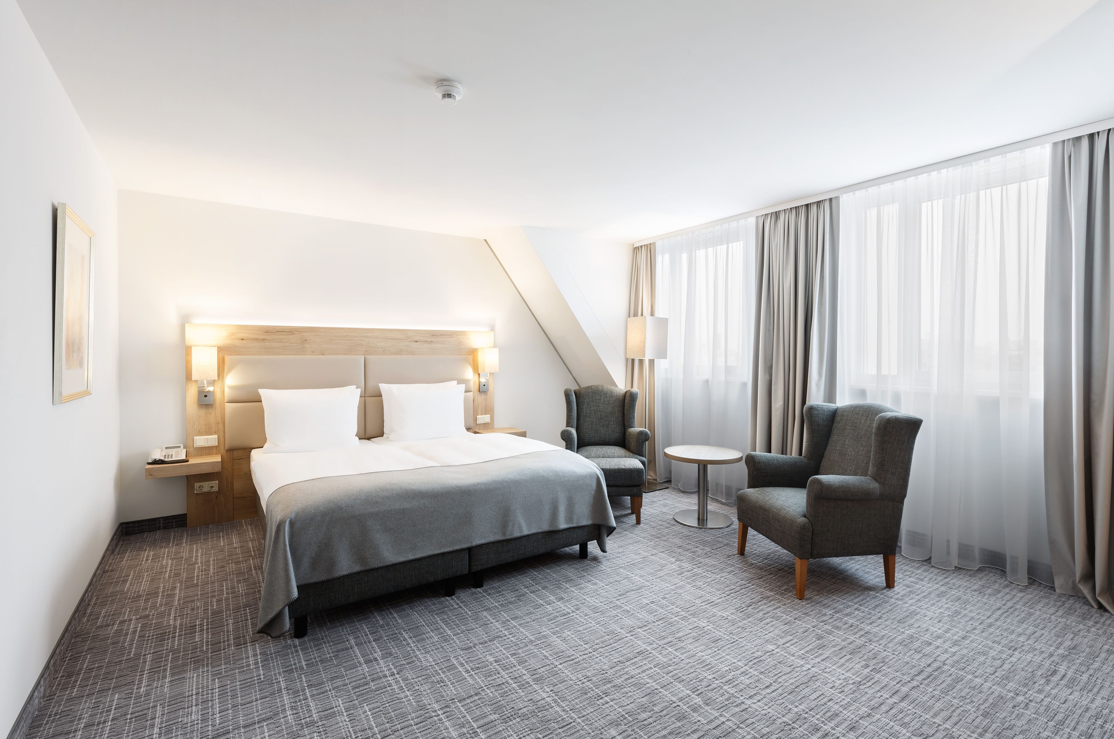 A hotel room with a bed, two armchairs, a small table, and a large window with curtains.