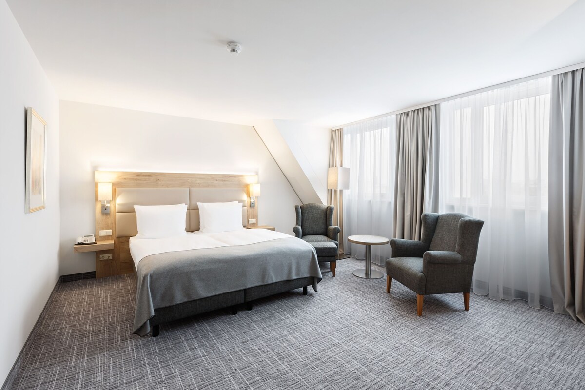 A hotel room with a bed, two armchairs, a small table, and a large window with curtains.