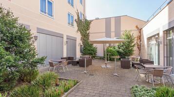 Ein Terrassenbereich im Freien mit Tischen und Stühlen, umgeben von Pflanzen und einer Gebäudefassade.