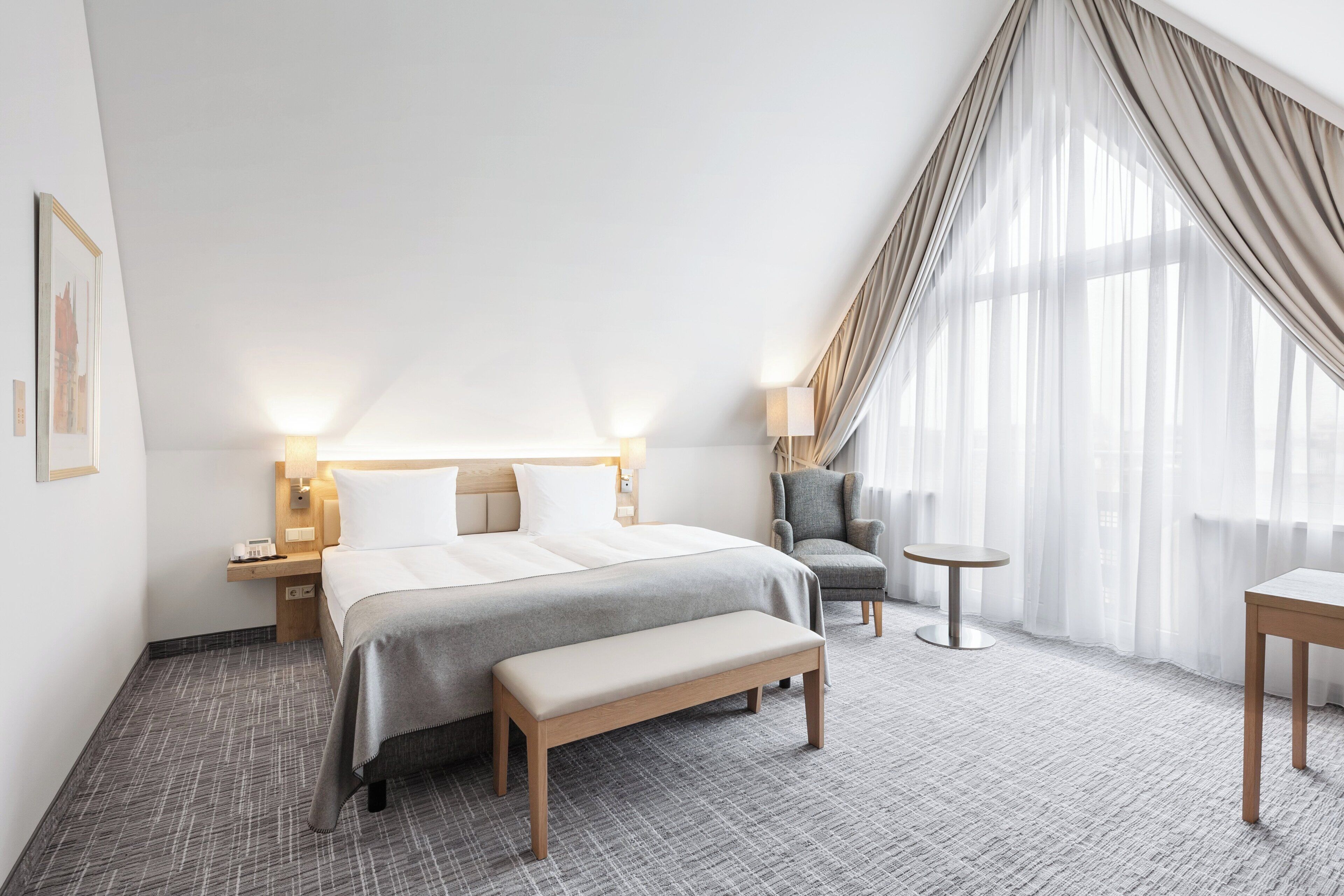 A hotel room with a bed, bedside tables, a chair, a small table, and a bench.