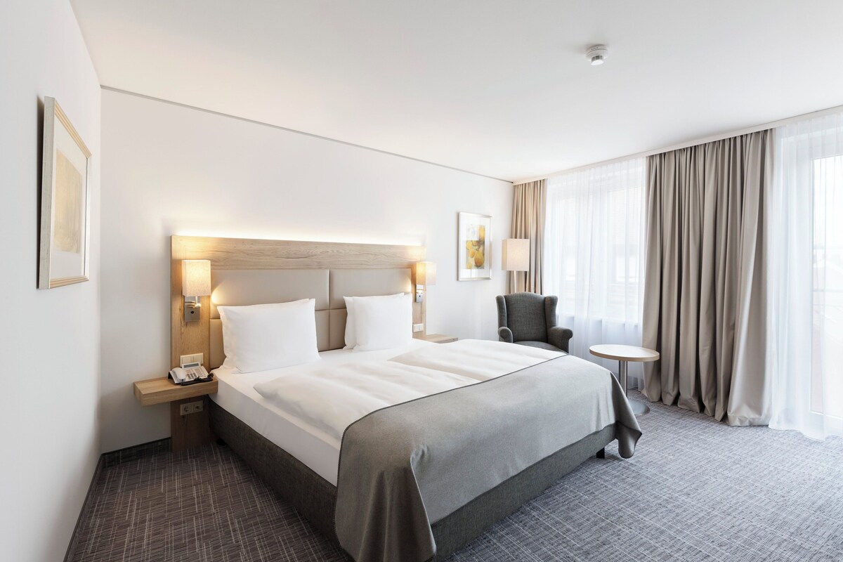 A hotel room with a large bed, bedside tables, a chair, a small round table, and a large window with curtains.