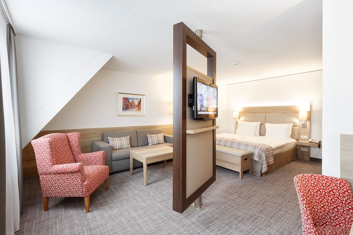A hotel room with a sofa, armchair, bed, TV, and bedside table.