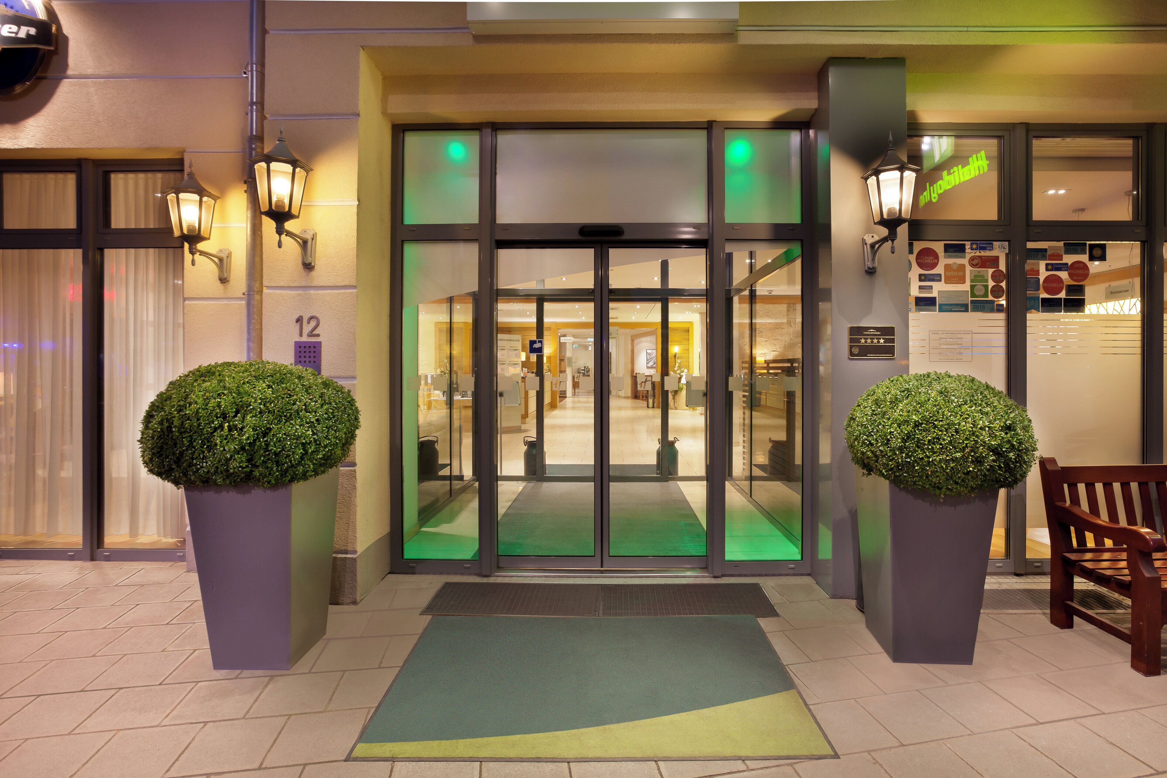 A hotel entrance with a glass door, a green mat, and two potted plants.