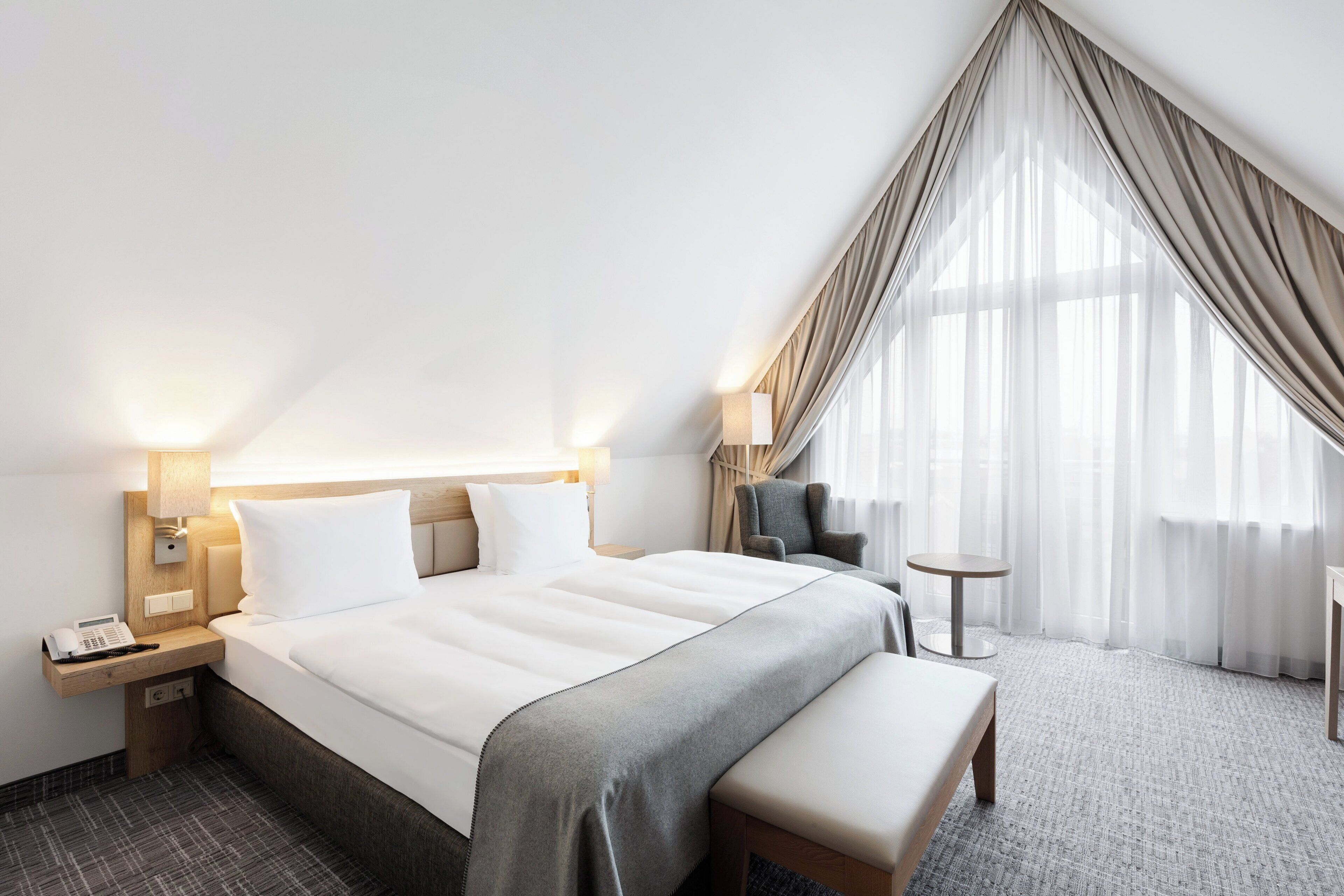 A hotel room with a large bed, bedside lamps, a small round table, a chair, and a bench.