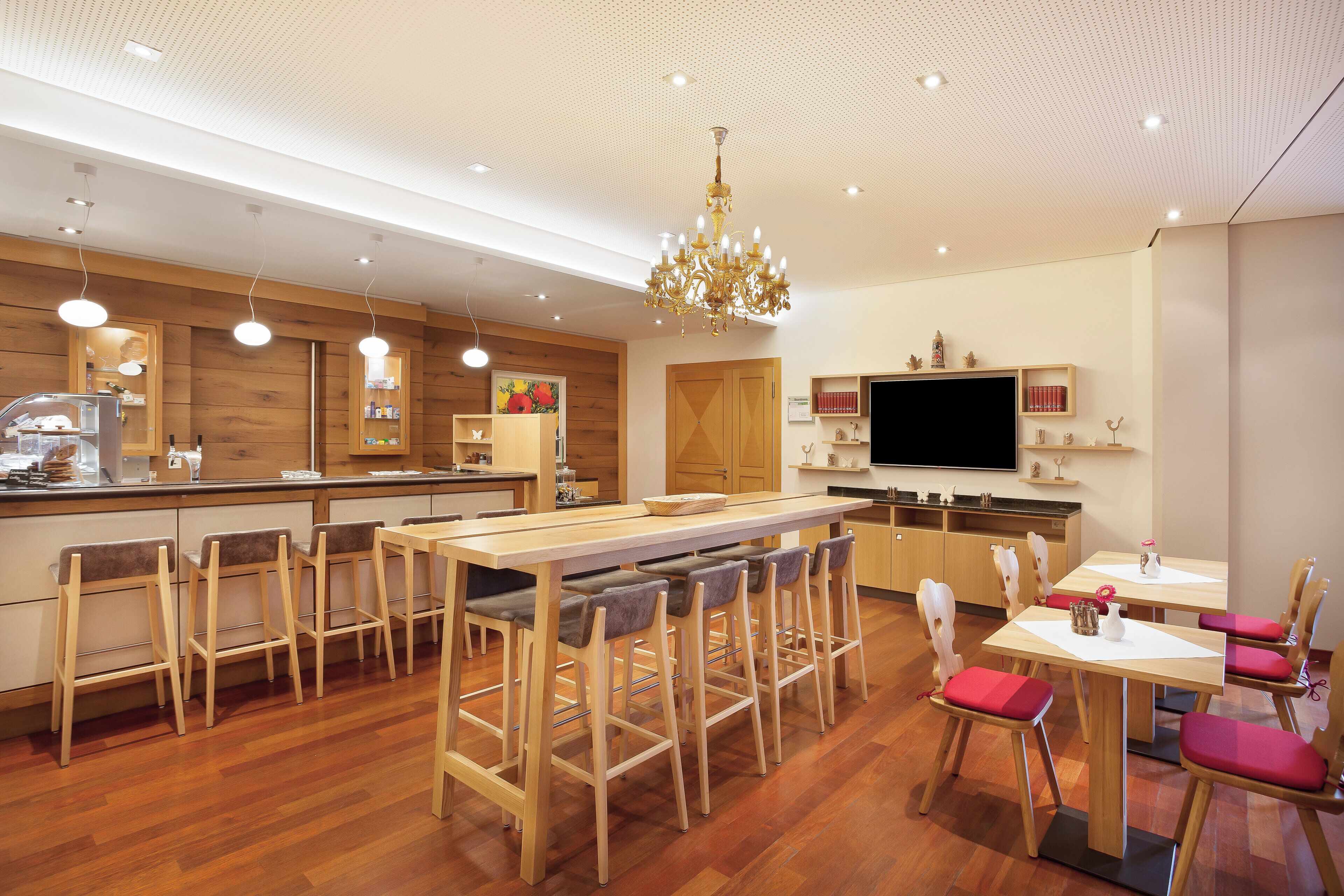 A modern kitchen with wooden cabinets, a central island with bar stools, a chandelier, and a flat-screen TV mounted on the wall.