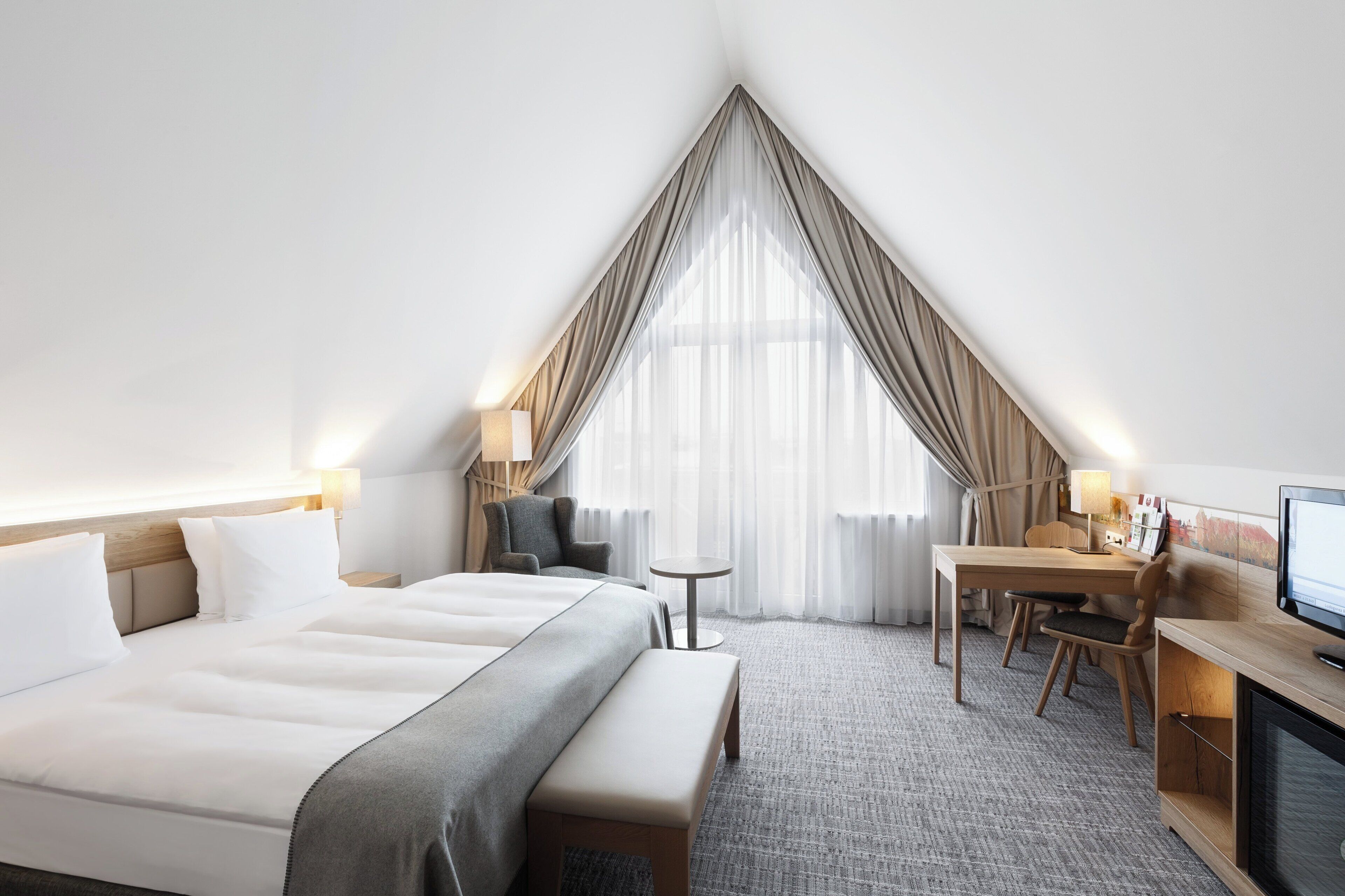 A hotel room with a large bed, a desk, a chair, a television, and a window with curtains.