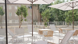 En udendørs terrasse med hvide stole og borde, parasoller og udsigt til en bygning med vinduer og træer.