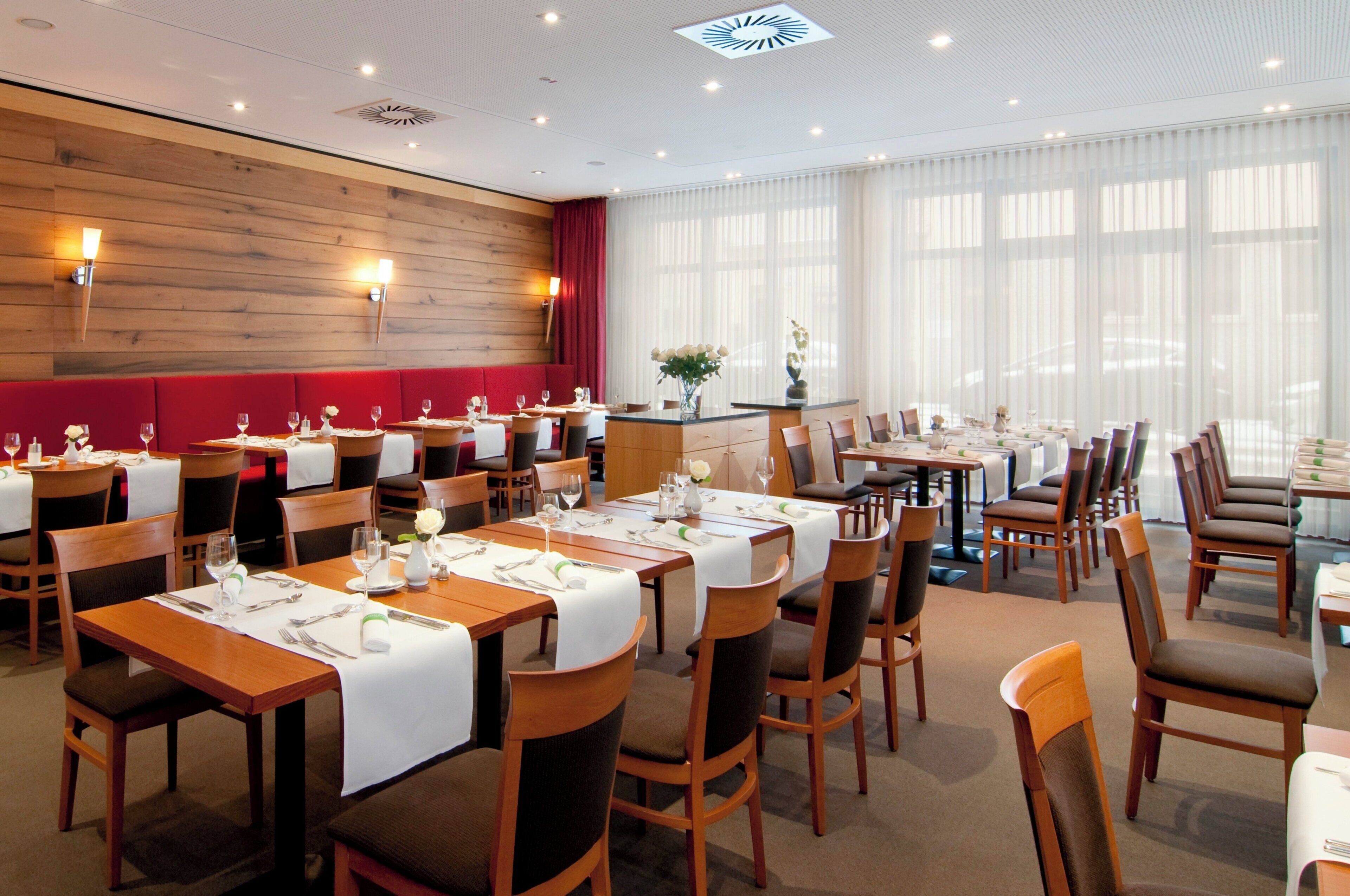 Comedor de hotel con varias mesas preparadas para comer, paredes de madera y ventanas grandes con cortinas translúcidas.