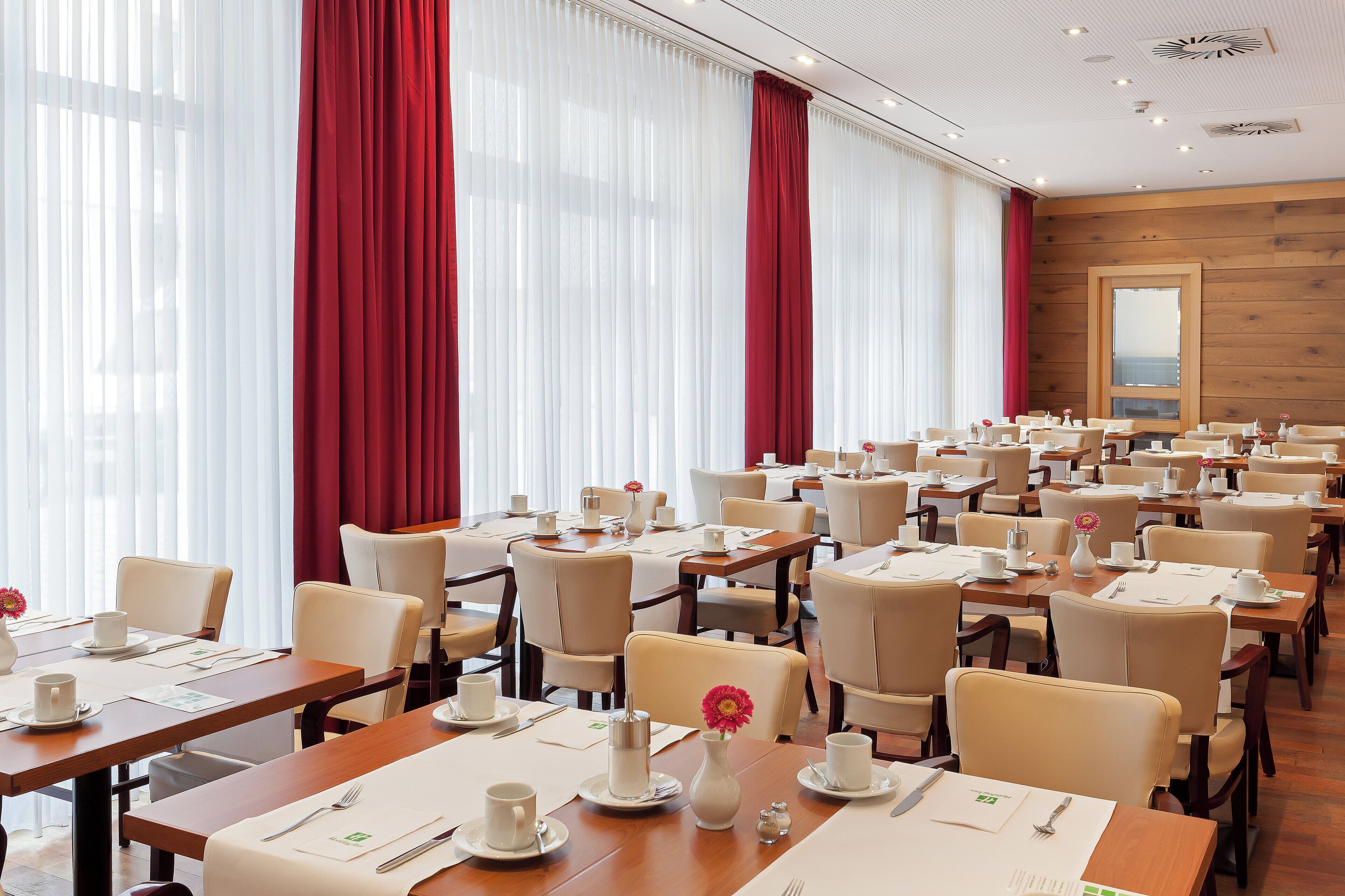 Comedor de hotel con varias mesas dispuestas para el desayuno, con manteles blancos, sillas y alimentos para el desayuno.
