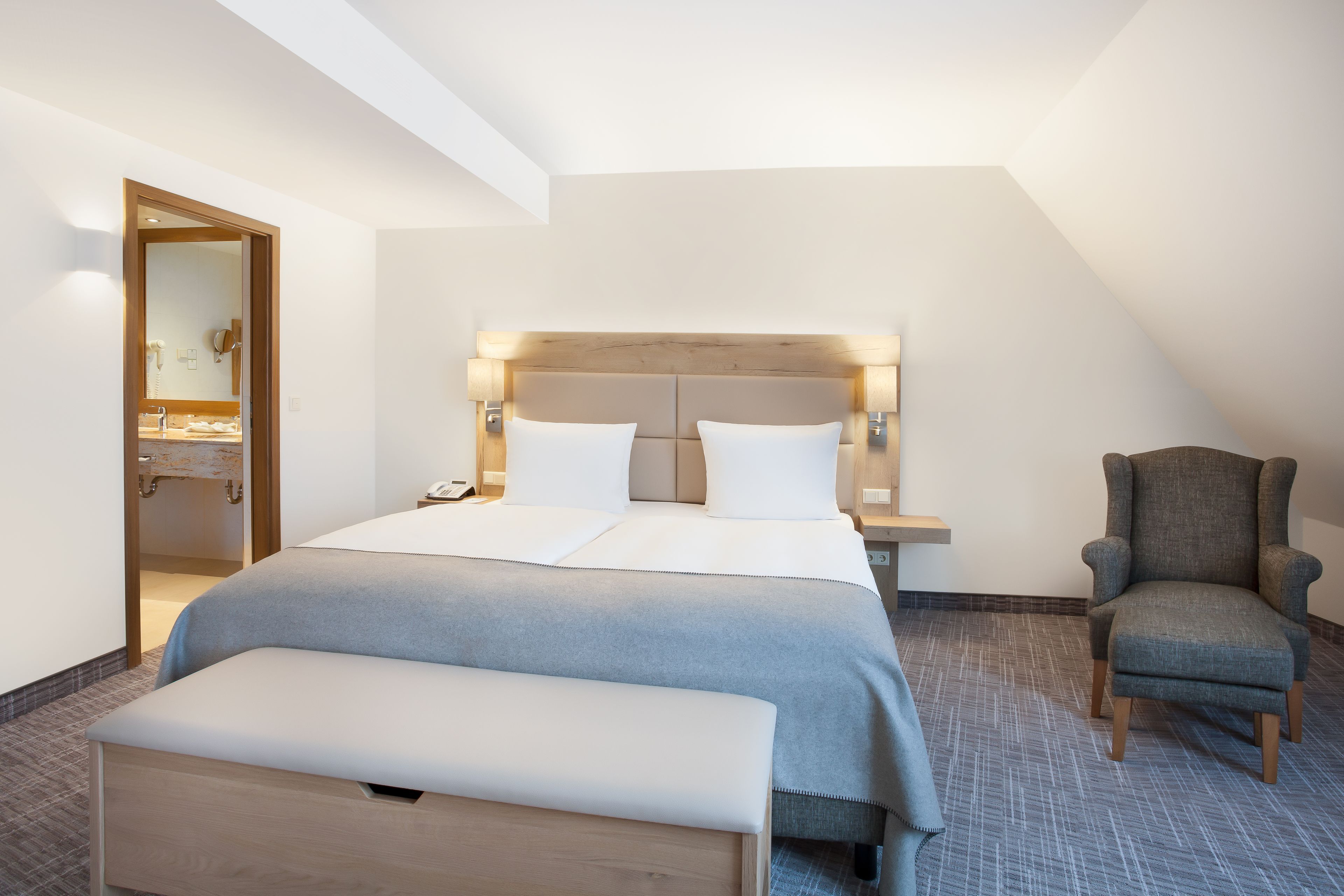 Une chambre d’hôtel moderne avec un grand lit, un banc, une chaise et une salle de bain visible à travers une porte ouverte.