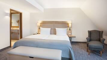 Una habitación de hotel moderna con una cama grande, un banco, una silla y un baño visible a través de una puerta abierta.