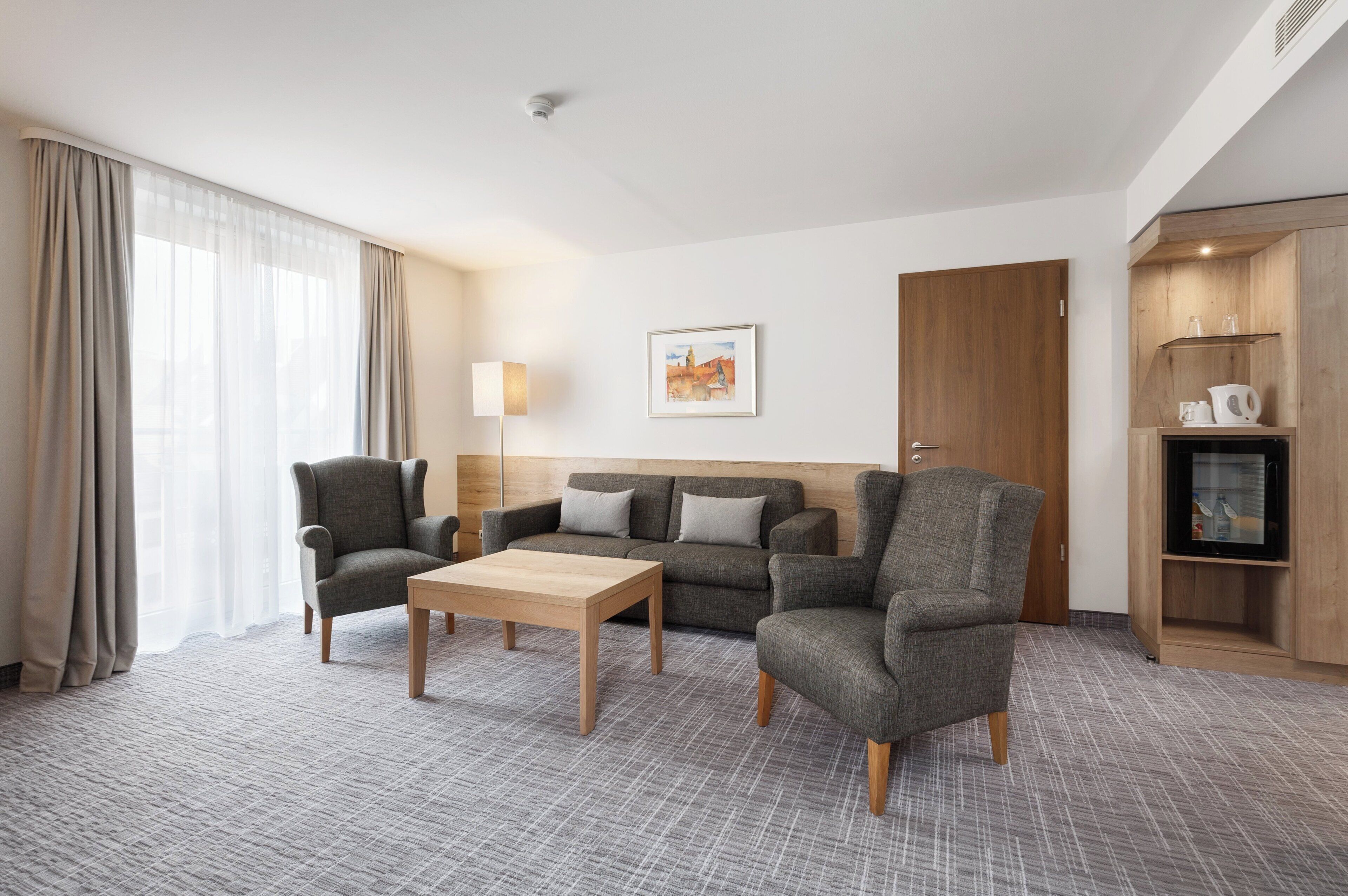 Habitación de hotel con sofá, dos sillones, una mesa de centro, una lámpara, un cuadro, un armario de madera y una puerta.