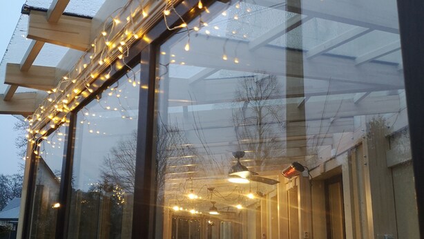 An outdoor dining area with wooden tables and chairs, string lights, and a glass enclosure with a ceiling fan. - Best Hotel (Riga)