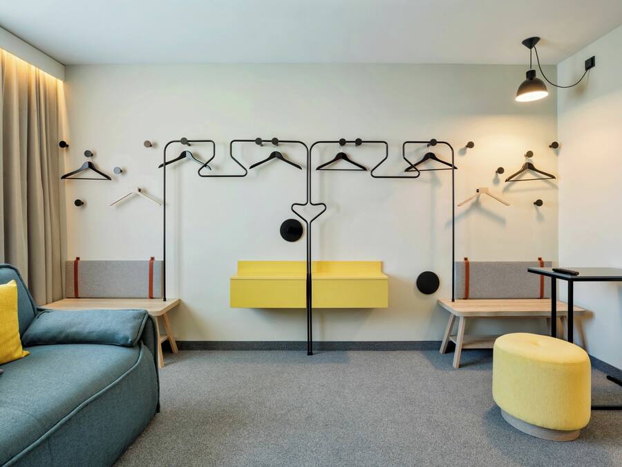 A modern hotel room with a sofa, ottoman, and a wall-mounted rack with multiple hangers.