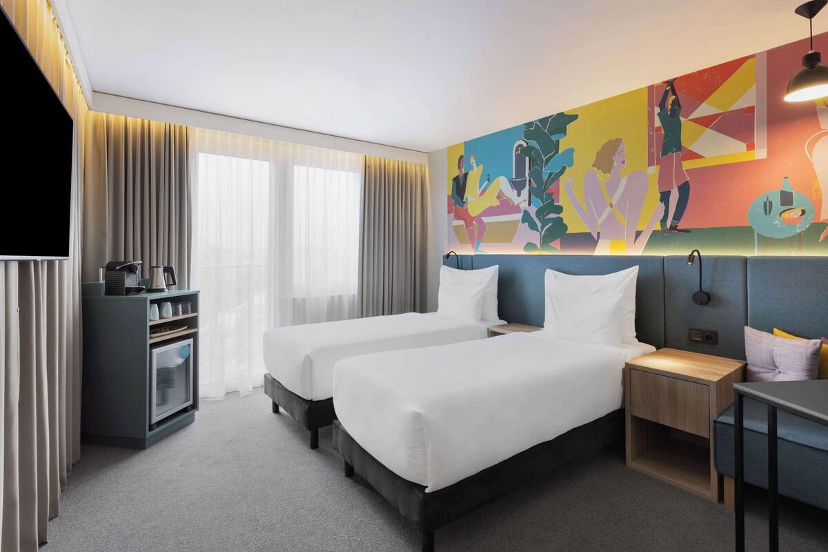 Ein Hotelzimmer mit zwei Betten, einem Fernseher, einem Schreibtisch und einem farbenfrohen Wandbild.