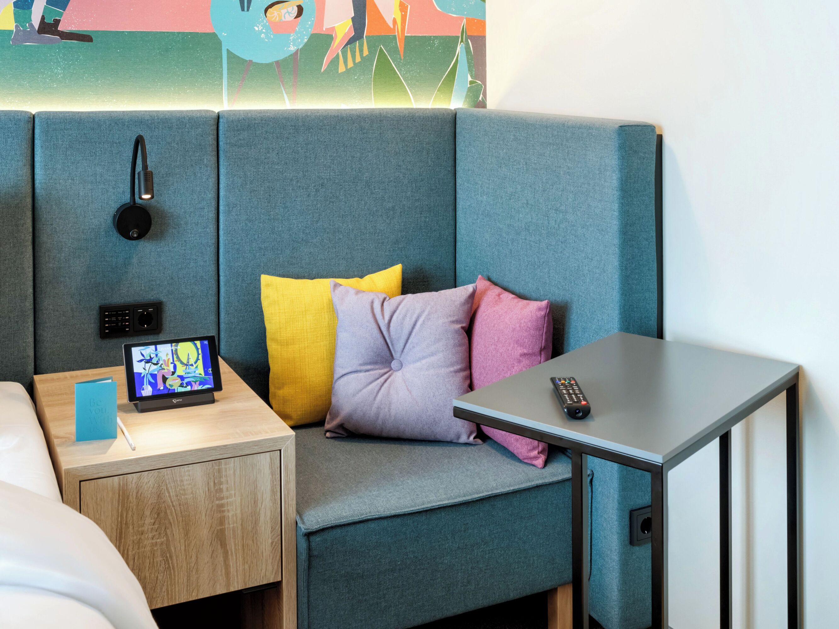 A modern hotel room with a grey sofa, a bedside table with a tablet, a remote control, and a wall-mounted speaker.