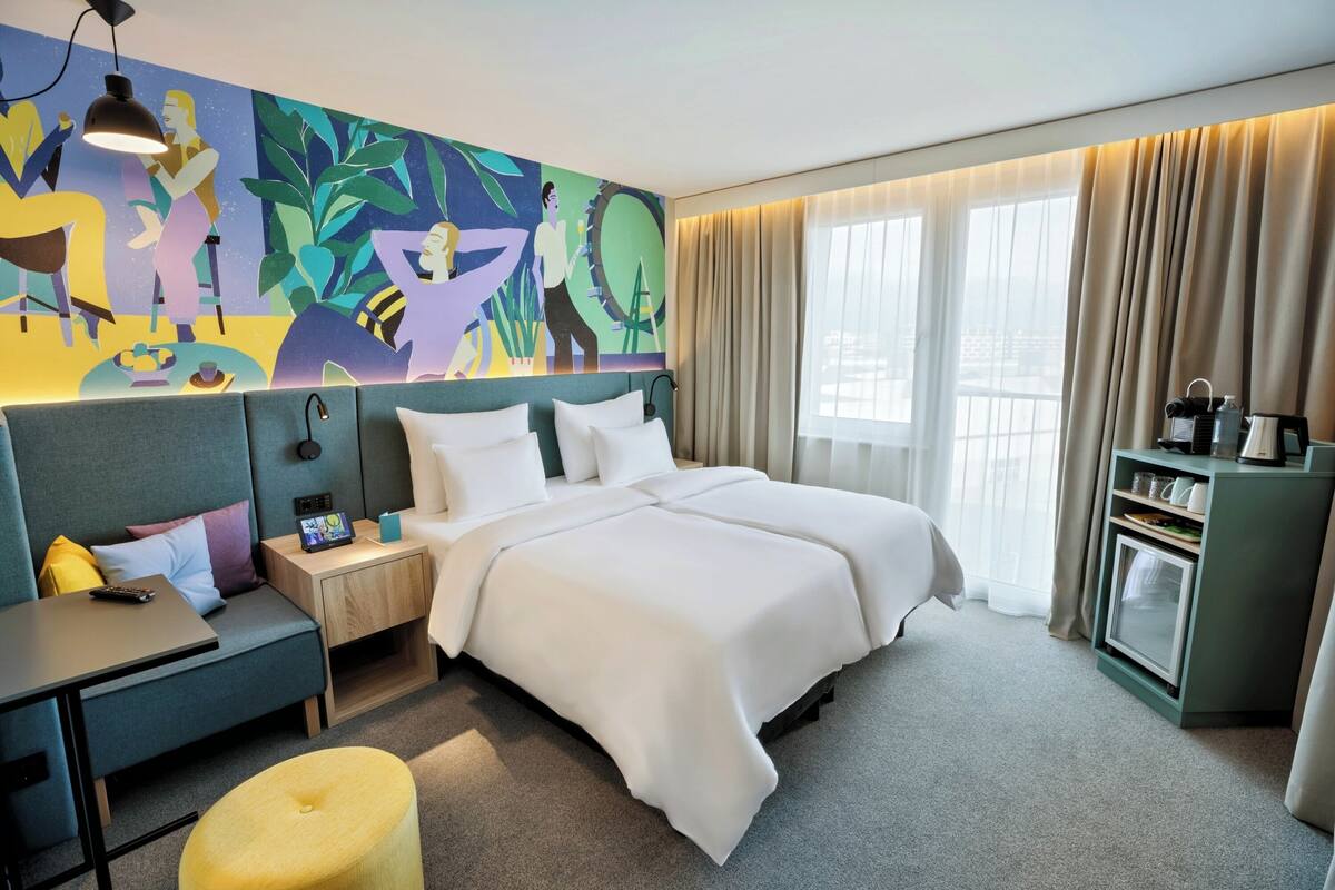 Ein Hotelzimmer mit einem großen Bett, einem Schreibtisch, einem kleinen Tisch, einem Nachttisch, Wandbildern, einem Fenster mit Vorhängen und einer Minibar.
