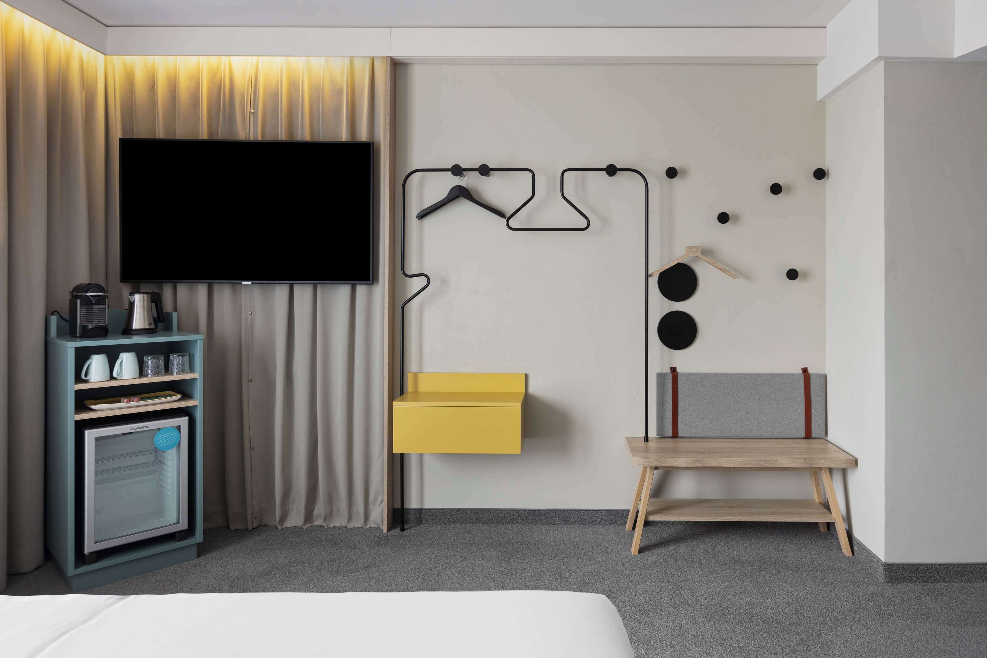 Hotellihuone, jossa on taulutelevisio, minibaari, keltainen säilytyslaatikko, puinen penkki ja seinään kiinnitetty naulakko.