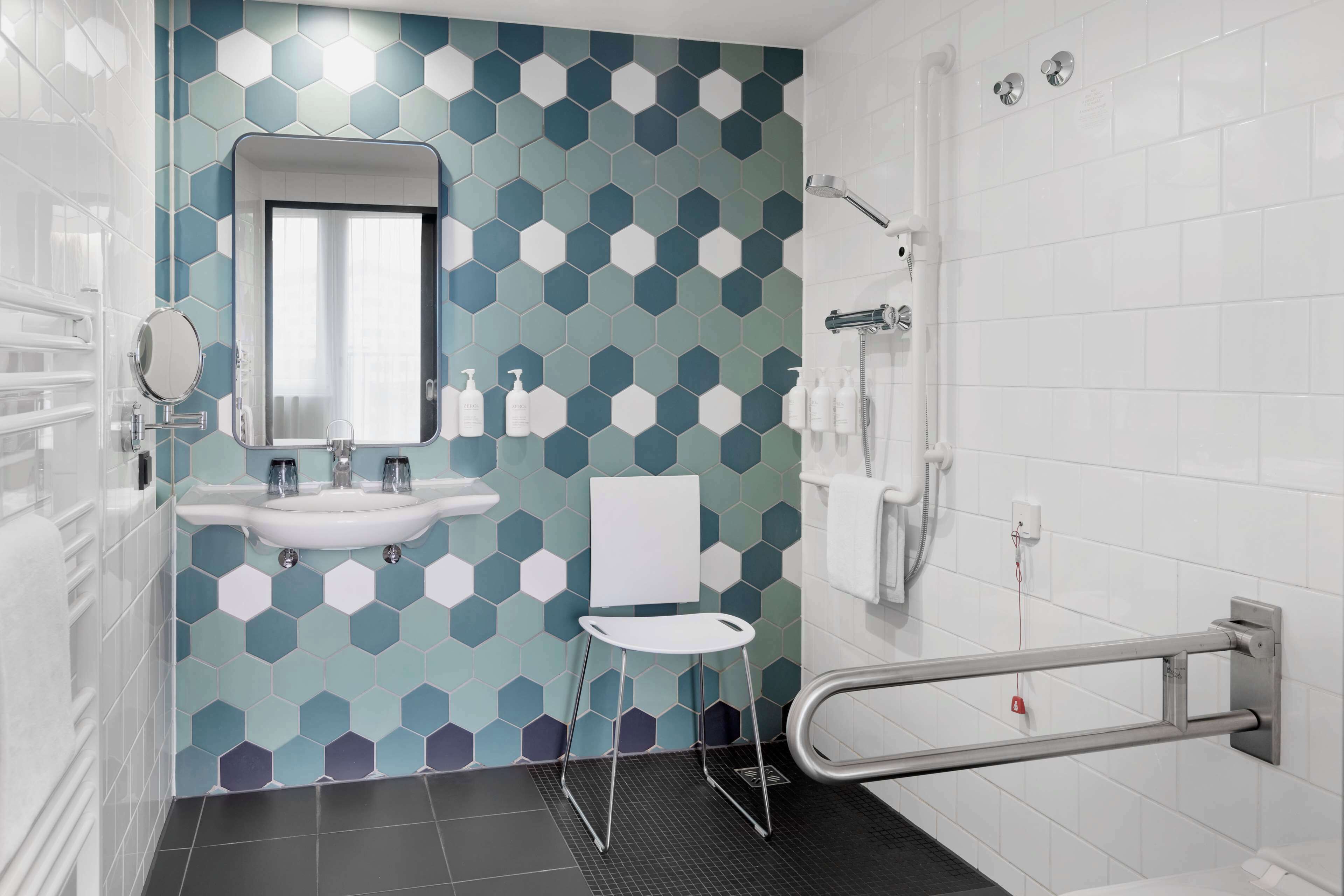 Un baño moderno con un salpicadero de azulejos hexagonales, un lavabo blanco, un espejo, una silla blanca, una zona de ducha y una barandilla.