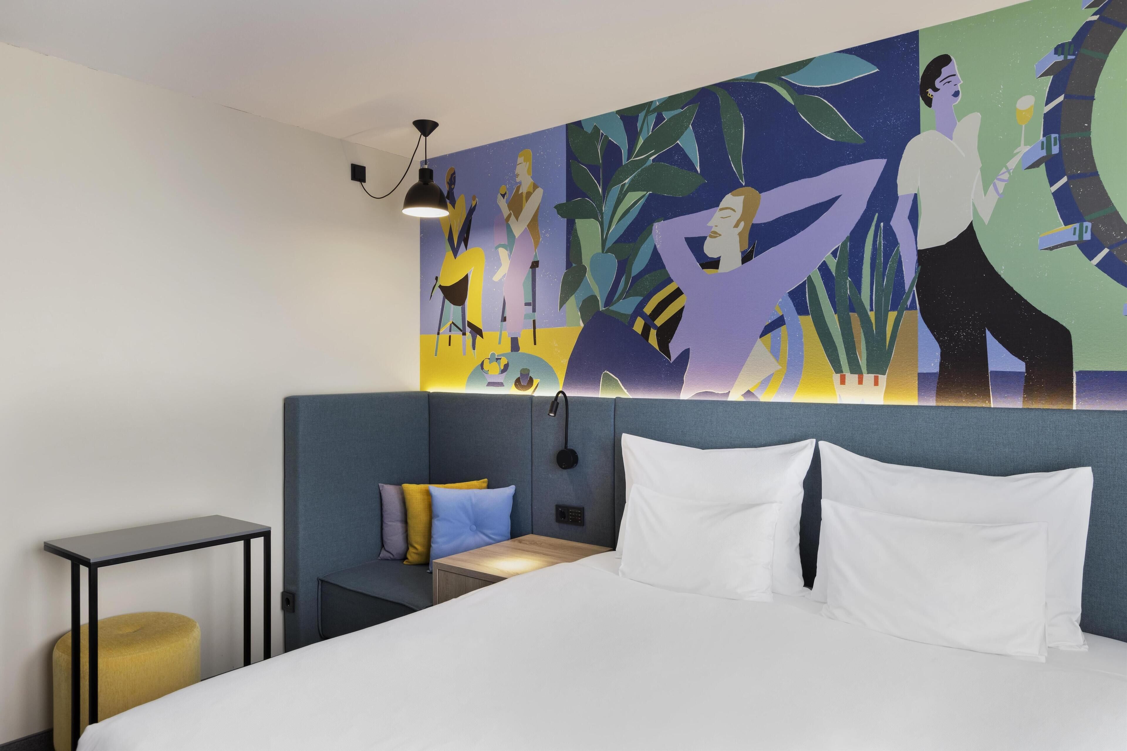 A modern hotel room with a large bed, a small bedside table, a nightstand, and a wall mural.