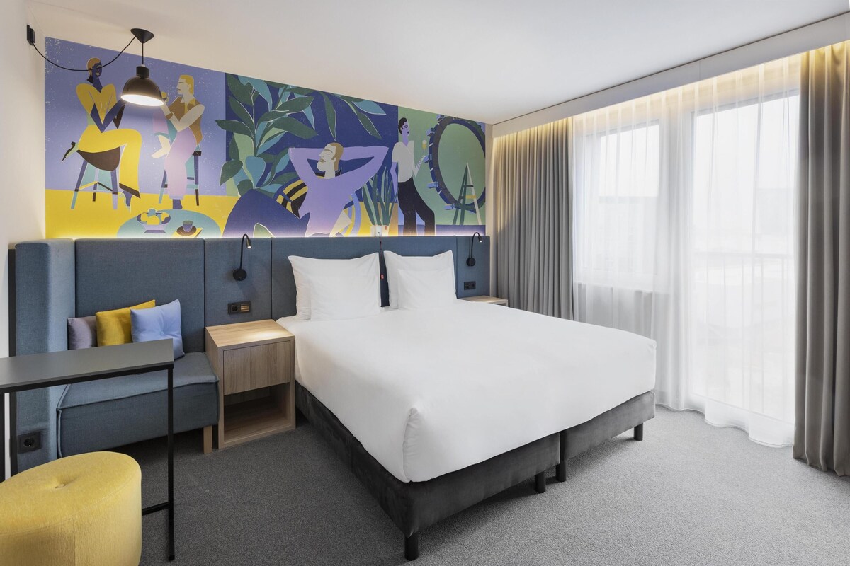 Een moderne hotelkamer met een groot bed, een bureau, een stoel en een kleurrijke abstracte muurschildering.
