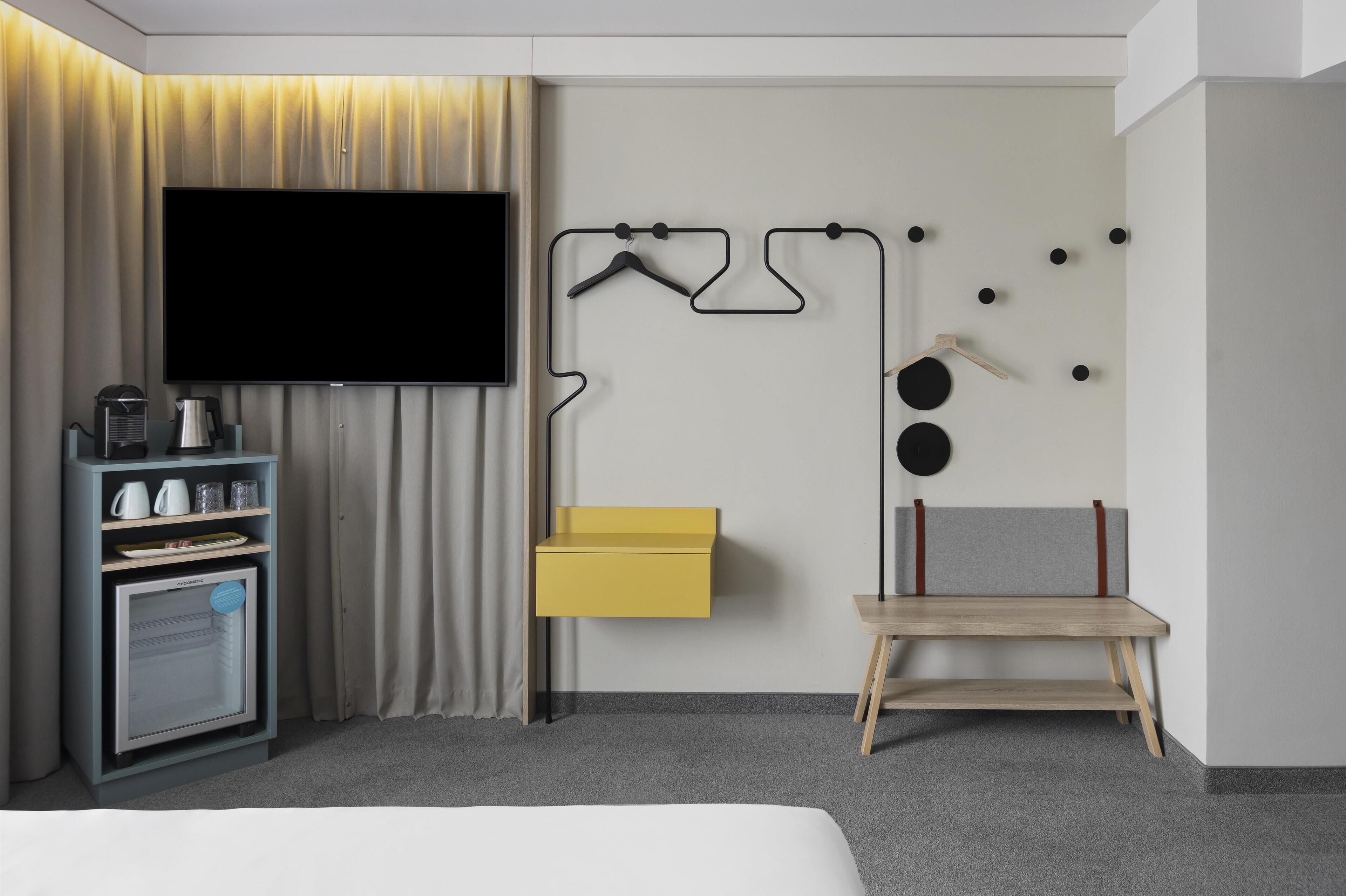 Une chambre d’hôtel équipée d’un téléviseur à écran plat, d’un mini-bar, d’un banc en bois et d’un porte-manteau mural.