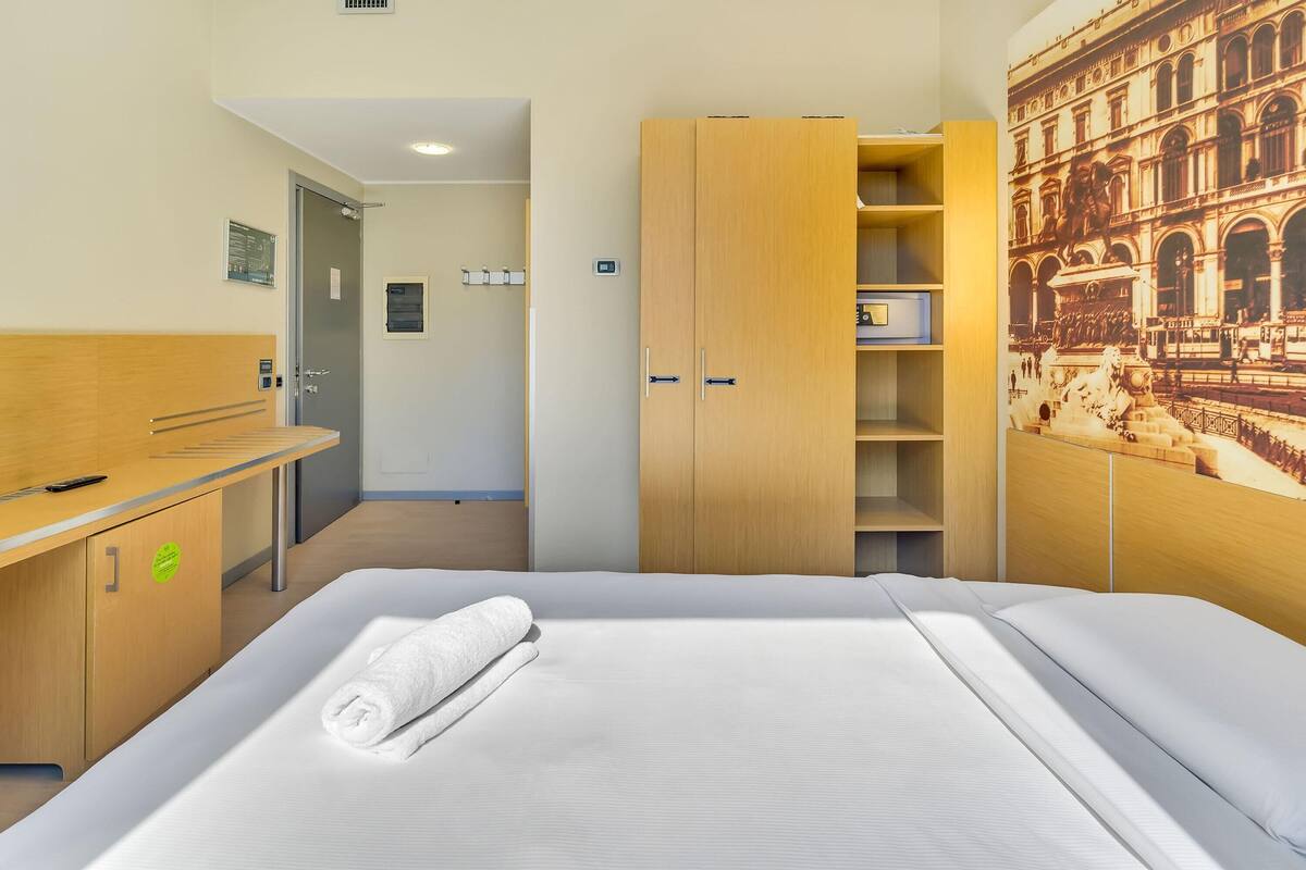 A hotel room with a bed, a wooden desk, a wardrobe, and a wall-mounted artwork.