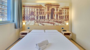 Una habitación de hotel con una cama grande, dos mesitas de noche, un cuadro colgado en la pared de un edificio histórico y una ventana con cortinas.