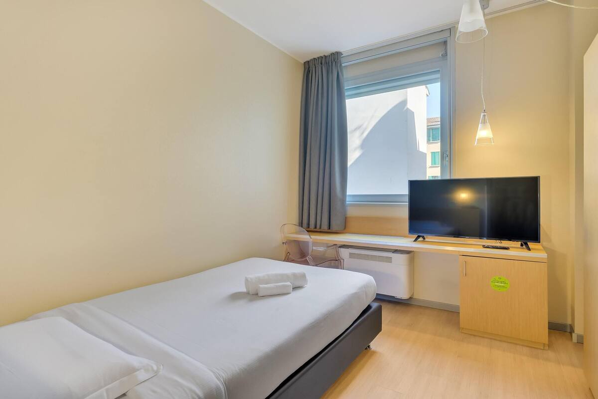 A hotel room with a bed, a desk with a TV, a window with curtains, and a cabinet.