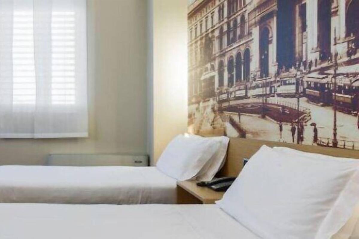 A hotel room with two beds, a sofa, a window with blinds, and a large cityscape painting on the wall.