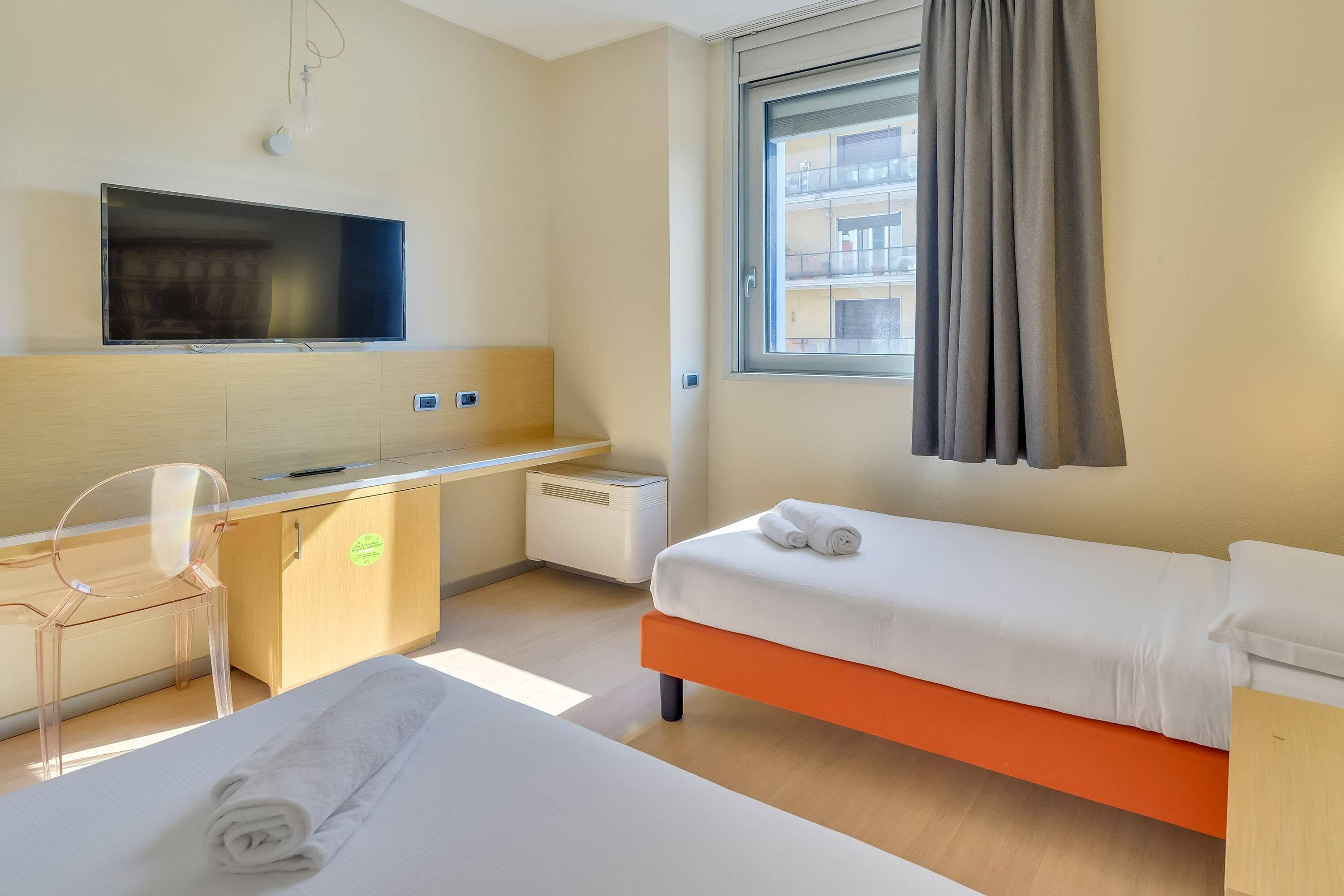 Una camera d'albergo con un letto, una scrivania, una sedia, una televisione e una finestra con tende.