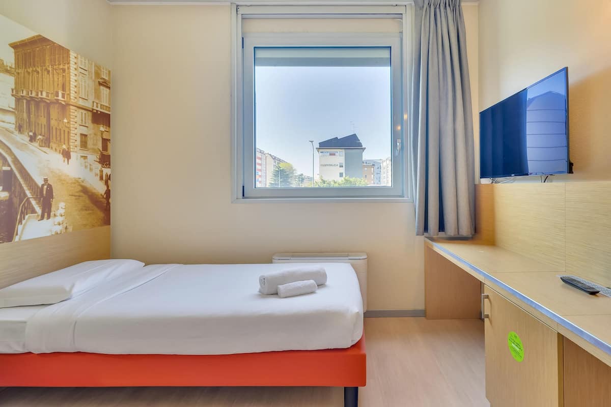 A hotel room with a bed, a window with a view of buildings, a TV mounted on the wall, and a wooden cabinet with a remote control on top.