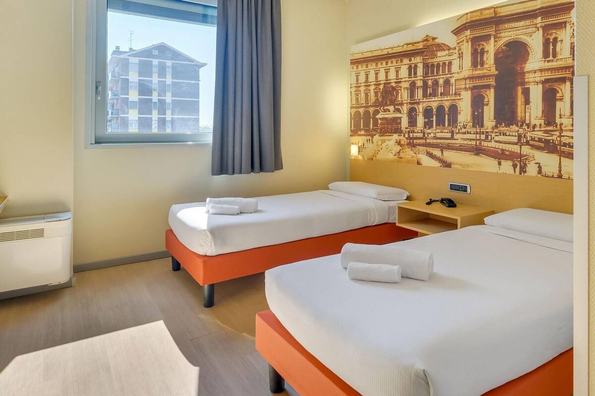 A hotel room with two beds, a window with curtains, a radiator, and a wall-mounted artwork.