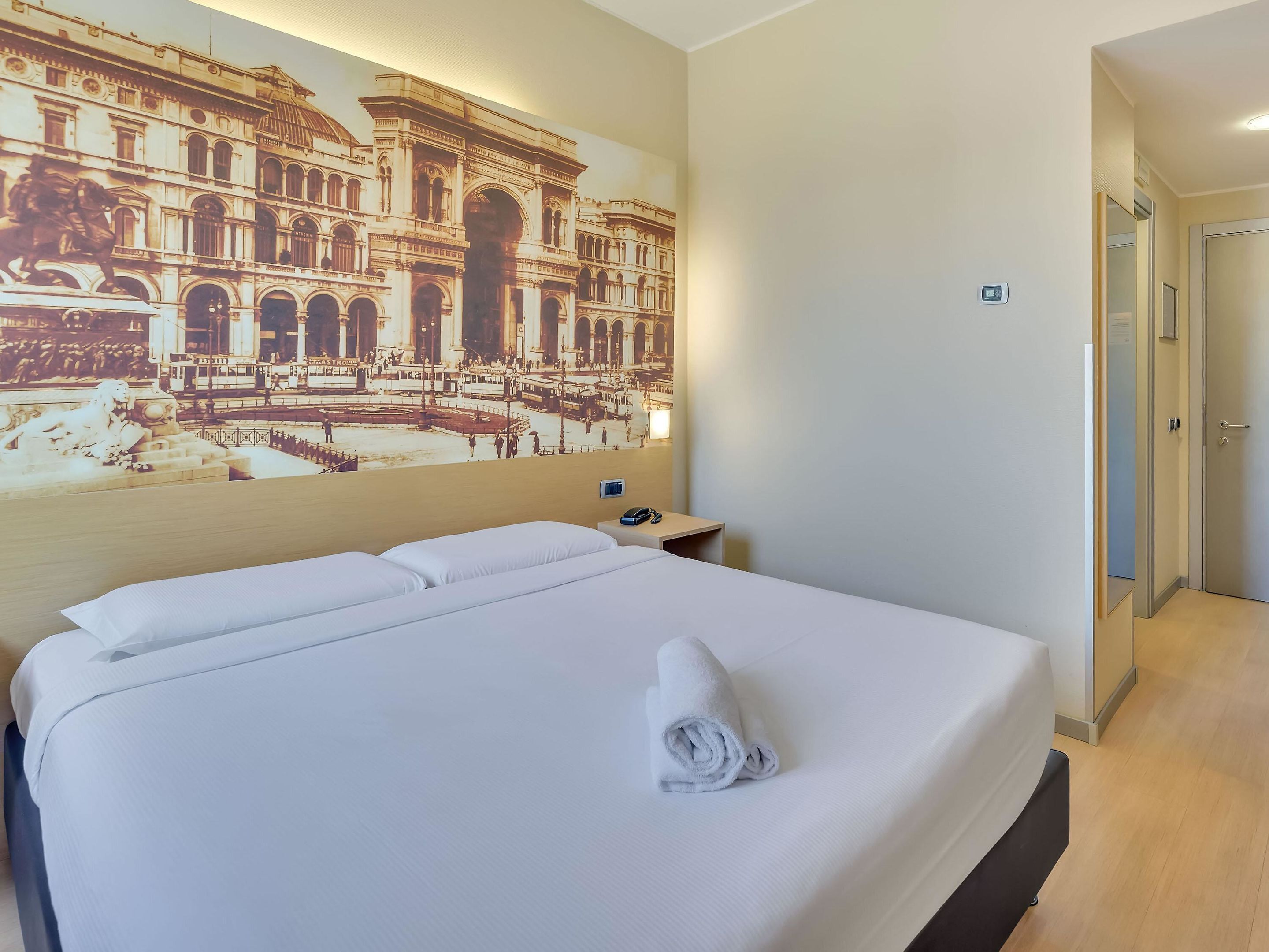 Ein Hotelzimmer mit einem großen Bett, einem Nachttisch, einer Wandlampe und Blick auf ein historisches Gebäude durch ein Fenster.
