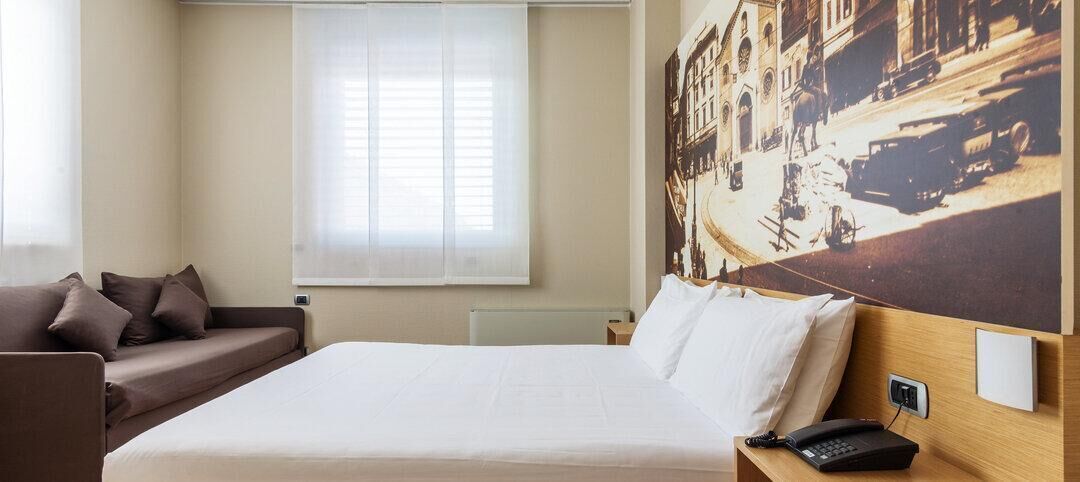 Een hotelkamer met een bed, een nachtkastje met telefoon, een raam met jaloezieën, een bank en een aan de muur hangend schilderij.