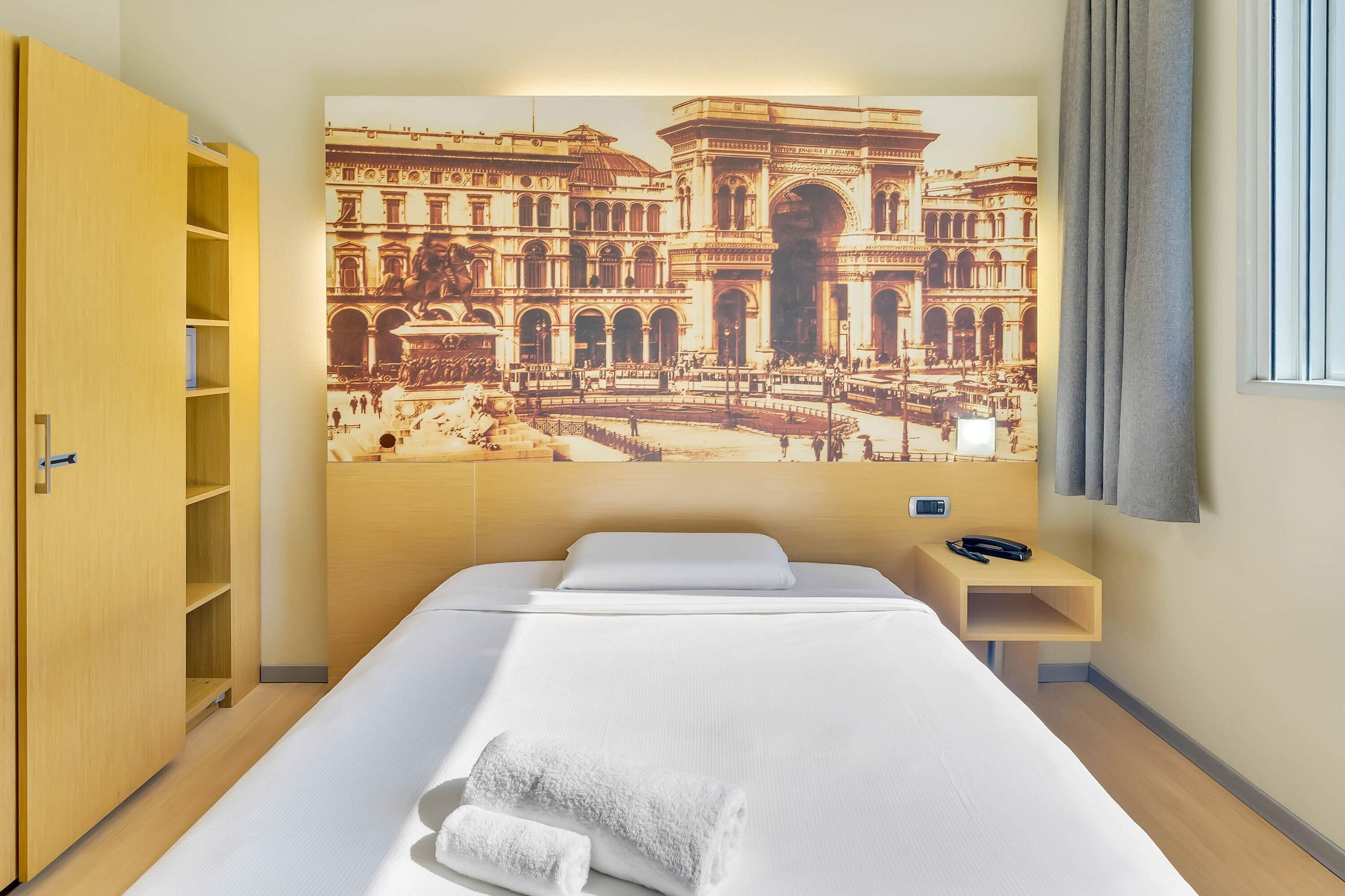 Ein Hotelzimmer mit einem Bett, einem Holzschrank, einem Nachttisch mit Telefon und einem großen Wandbild eines historischen Gebäudes.