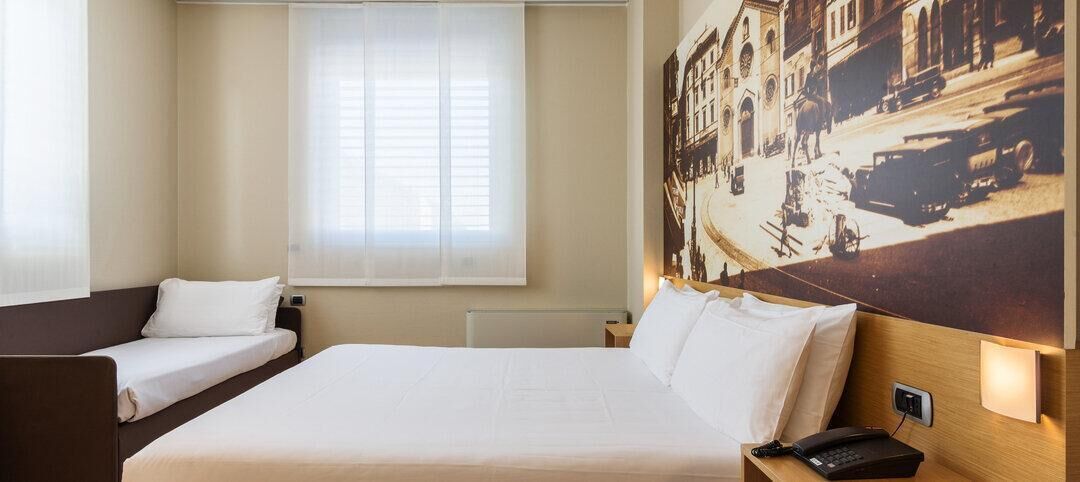 Ein Hotelzimmer mit zwei Einzelbetten, einem großen Fenster mit leichtem Vorhang, einem wandmontierten Telefon und einem Vintage-Stadtbild.
