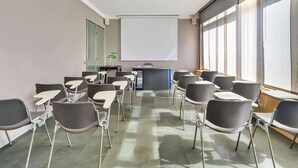 Un salón de conferencias con filas de sillas, un atril, una pantalla de proyector y amplias ventanas.