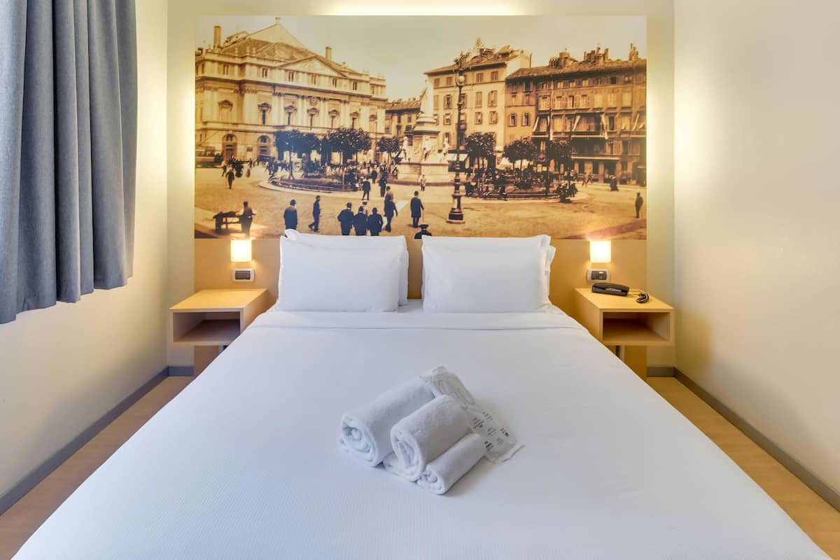 A hotel room with a large bed, two pillows, a neatly folded towel, and a wall mural depicting a historical scene.