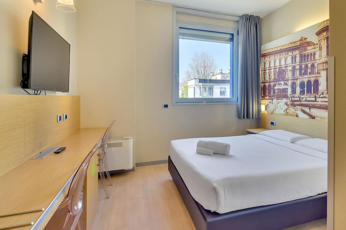 A modern hotel room with a bed, a TV mounted on the wall, a window with a view of buildings, and a wall-mounted air conditioner.