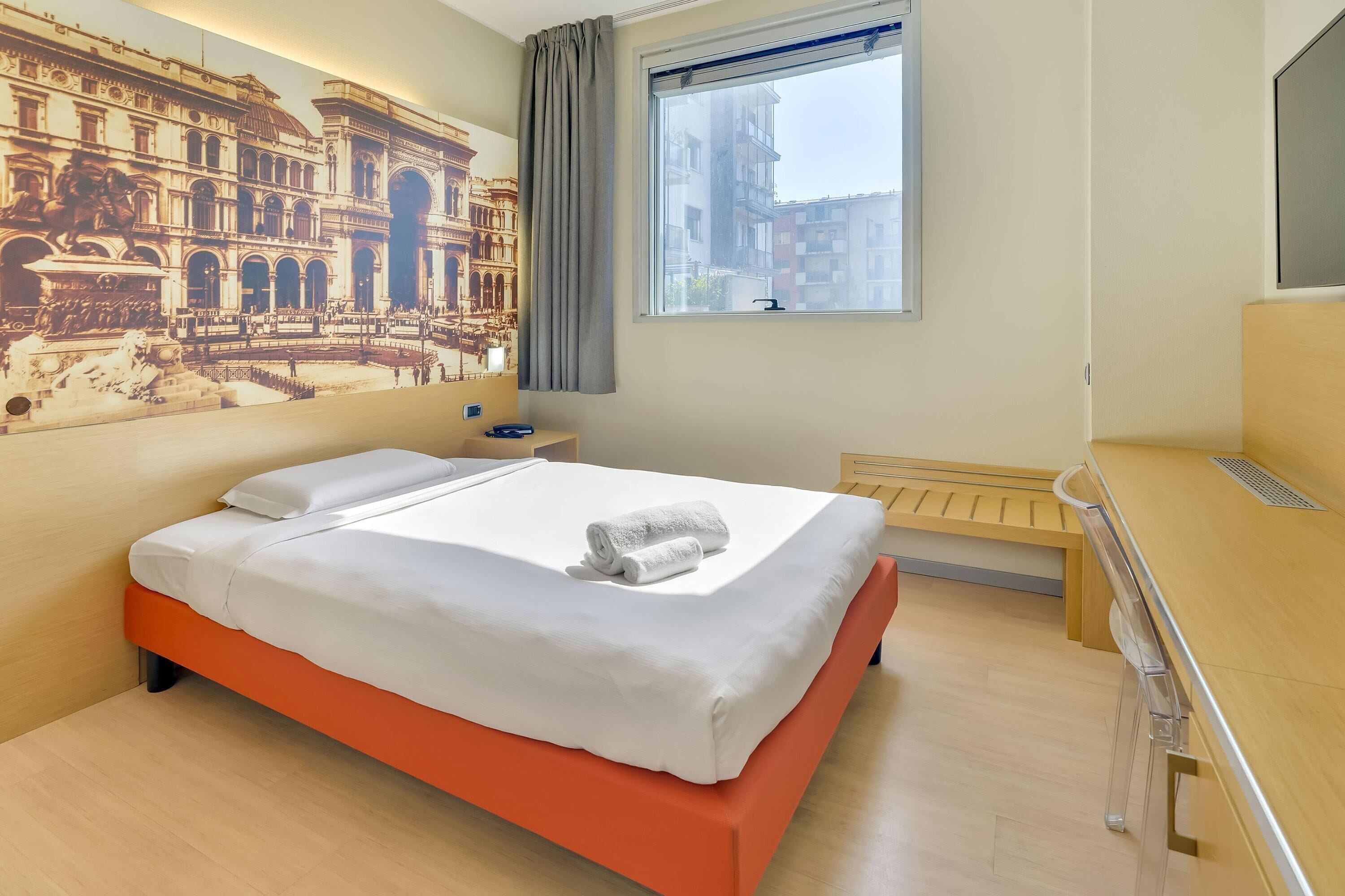 A hotel room with a bed, a desk, a window with a view of buildings, and a mural on the wall.