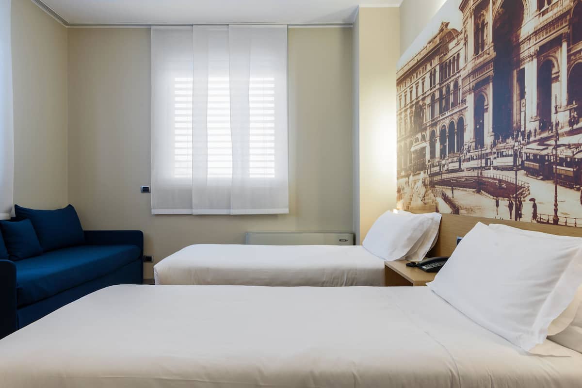 A hotel room with two beds, a blue sofa, a window with blinds, and a large historical building mural on the wall.