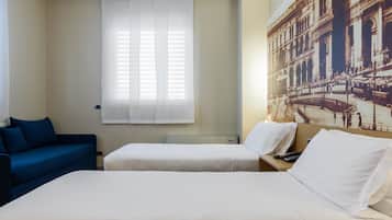 Camera d'albergo con due letti, un divano blu, una finestra con persiane e un grande murale raffigurante un edificio storico.