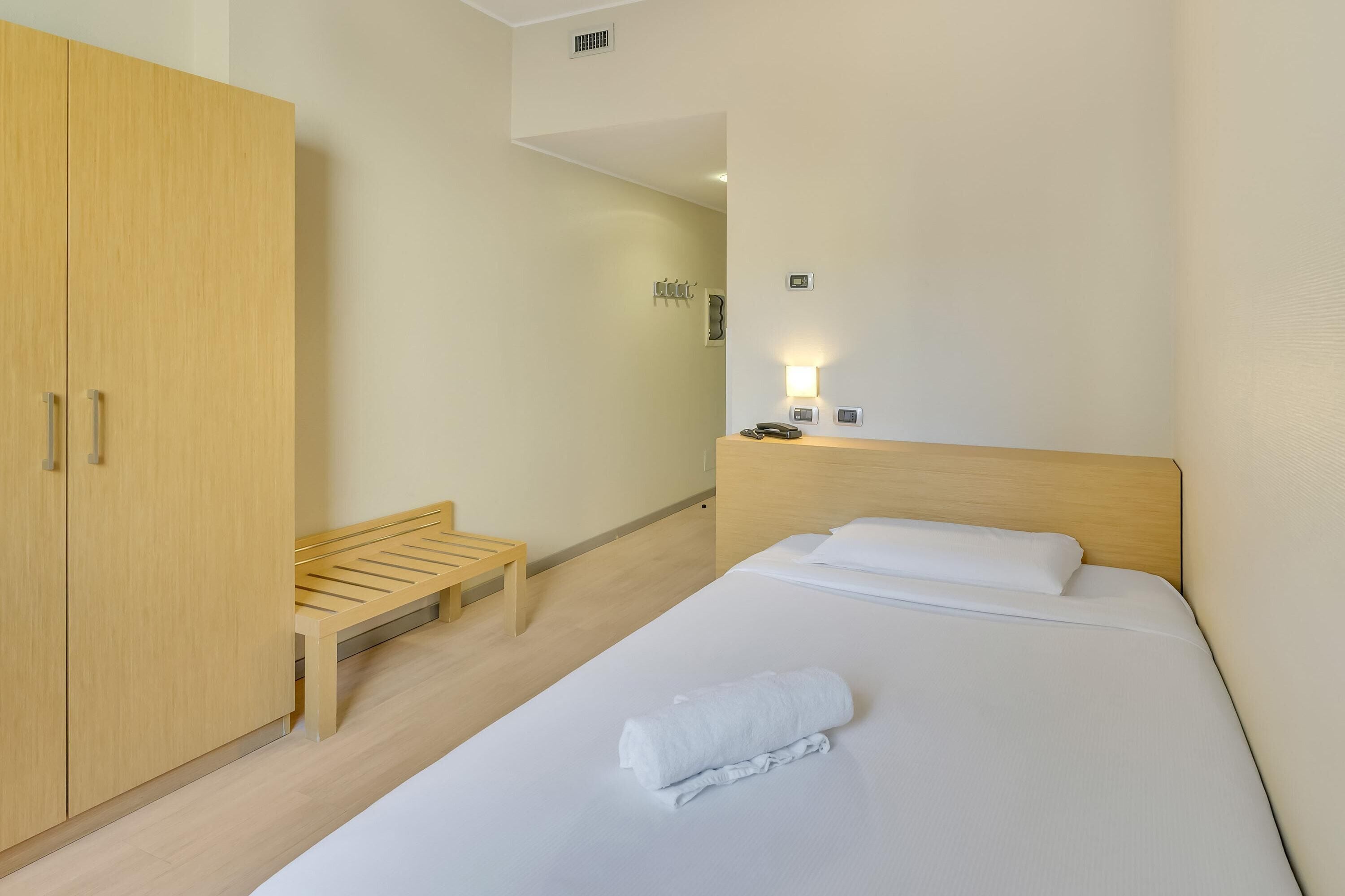 Une chambre d’hôtel avec un lit, une armoire en bois, un banc et une table de chevet avec un téléphone et une lampe.
