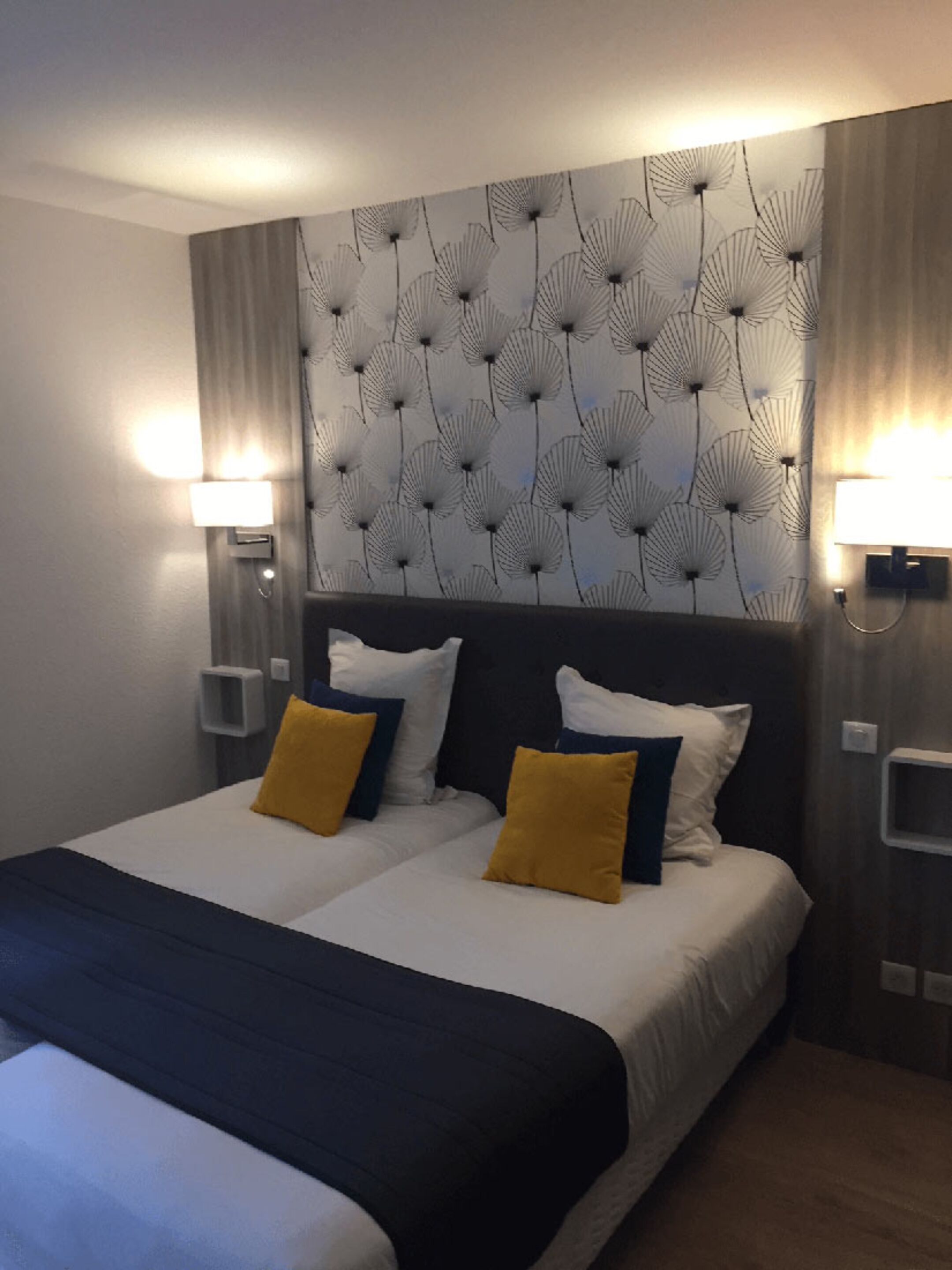 A double bed with white and black bedding, two yellow pillows, and a dark headboard. There are two wall-mounted lamps on either side of the bed.