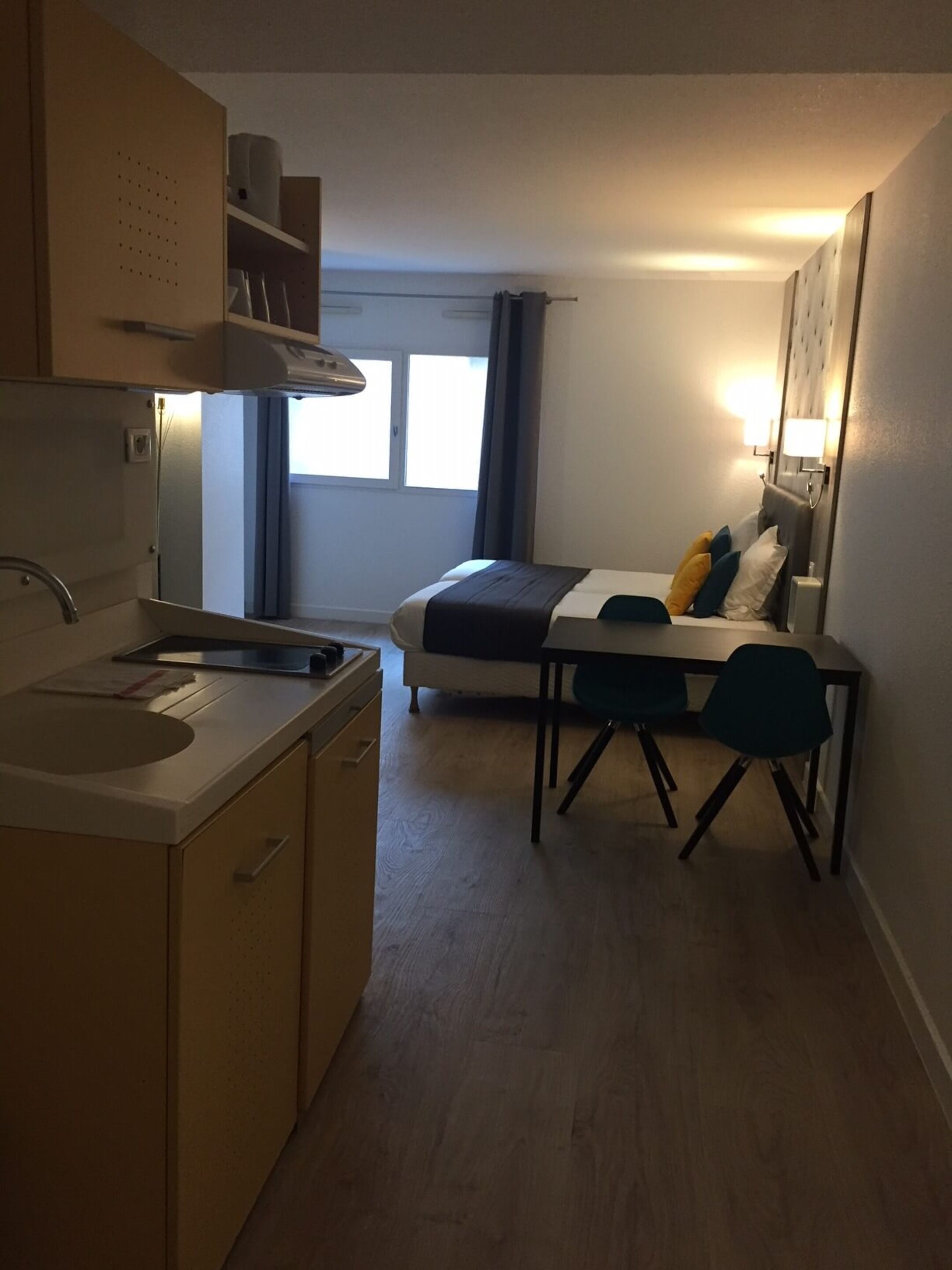 A compact hotel room with a kitchenette, a bed with pillows, a small table, and a chair.