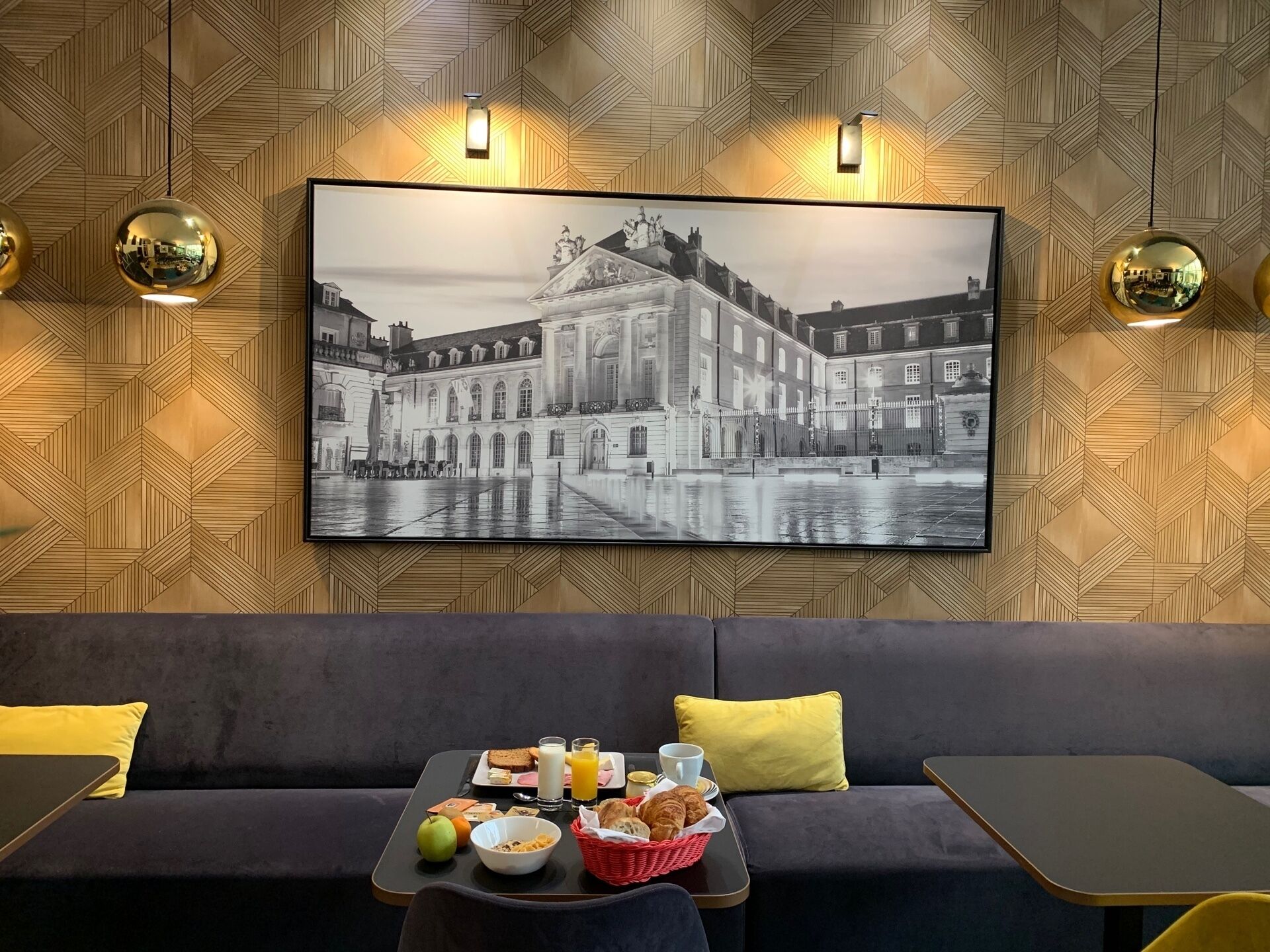 Une chambre d’hôtel moderne, avec une grande photographie en noir et blanc encadrée représentant un bâtiment historique accrochée au mur, un canapé sombre agrémenté de coussins jaunes et un plateau de petit-déjeuner posé sur une petite table.