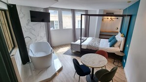 Un dormitorio moderno con una cama grande, una zona de estar con una mesa y sillas, una bañera y un televisor montado en la pared.