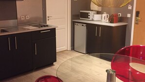 Una cocina moderna con armarios oscuros, una estufa de acero inoxidable, un microondas y un hervidor rojo. Hay una mesa de cristal con sillas rojas y una lámpara colgante original.